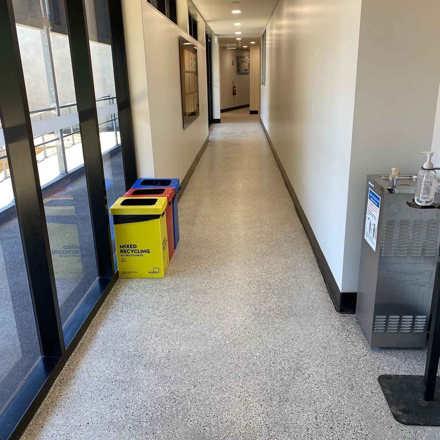 Long glazed corridor with an even speckled floor and recycling bins